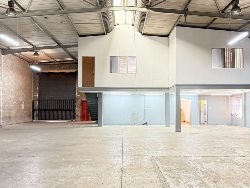 Spacious industrial warehouse featuring a mezzanine level, high ceilings with exposed steel trusses, concrete flooring, and a large roller shutter door for easy access.
