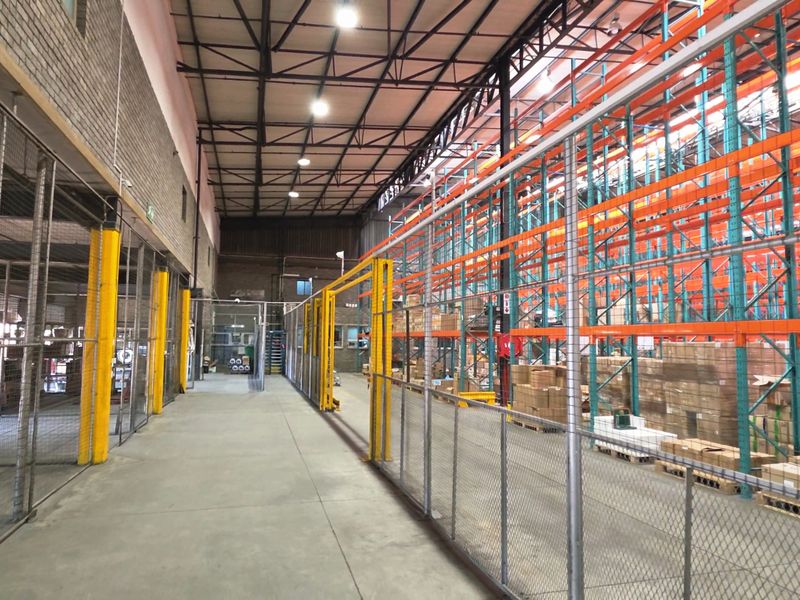Spacious industrial warehouse featuring high-clearance steel roof trusses, concrete flooring, and extensive heavy-duty steel racking systems with secure wire mesh partitioning.