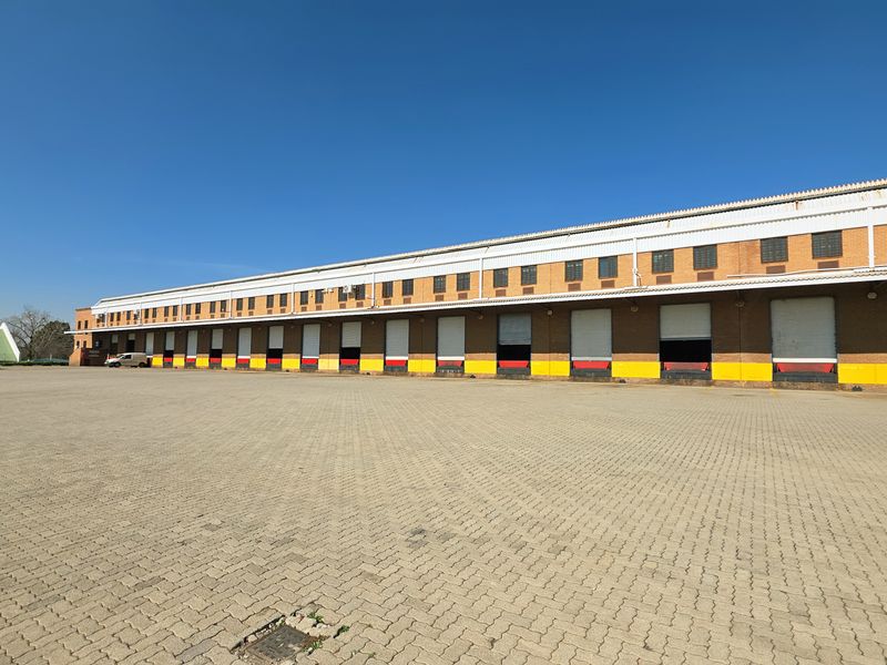 Large industrial warehouse facility featuring multiple loading bays with roller shutter doors and an expansive paved yard area for logistics and transport access.