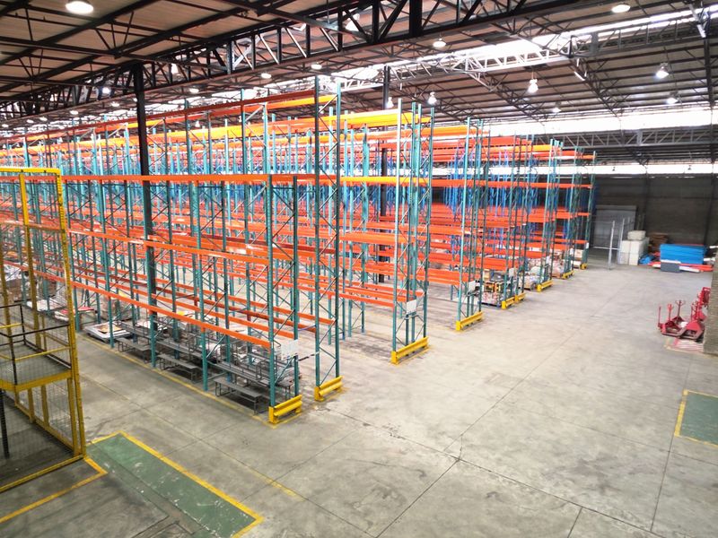 Spacious industrial warehouse featuring high-bay lighting, exposed steel roof trusses, concrete flooring, and an extensive integrated racking system for storage.