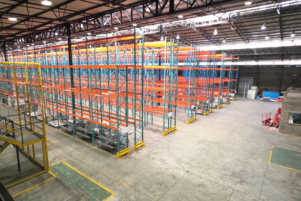 Spacious industrial warehouse featuring high-bay lighting, exposed steel roof trusses, concrete flooring, and an extensive integrated racking system for storage.