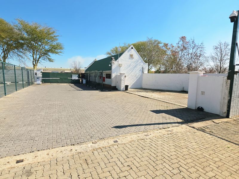 Industrial property entrance featuring a paved driveway, secure perimeter fencing, and gated access point with security infrastructure.