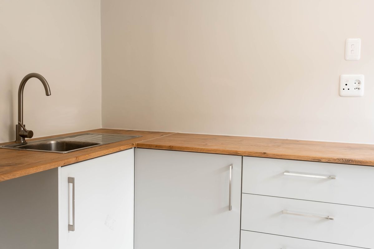 A clean, minimalist kitchen area featuring light grey cabinetry, a wooden countertop, and a modern stainless steel sink with a gooseneck faucet.
