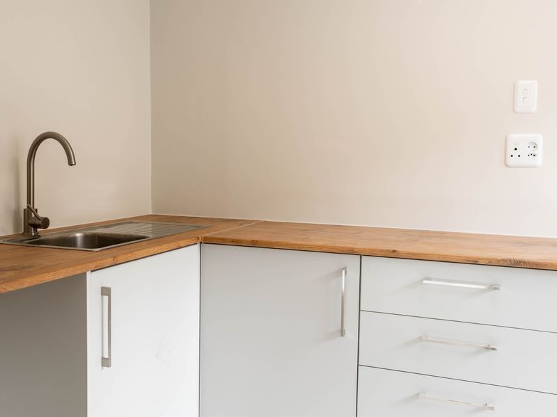 A clean, minimalist kitchen area featuring light grey cabinetry, a wooden countertop, and a modern stainless steel sink with a gooseneck faucet.