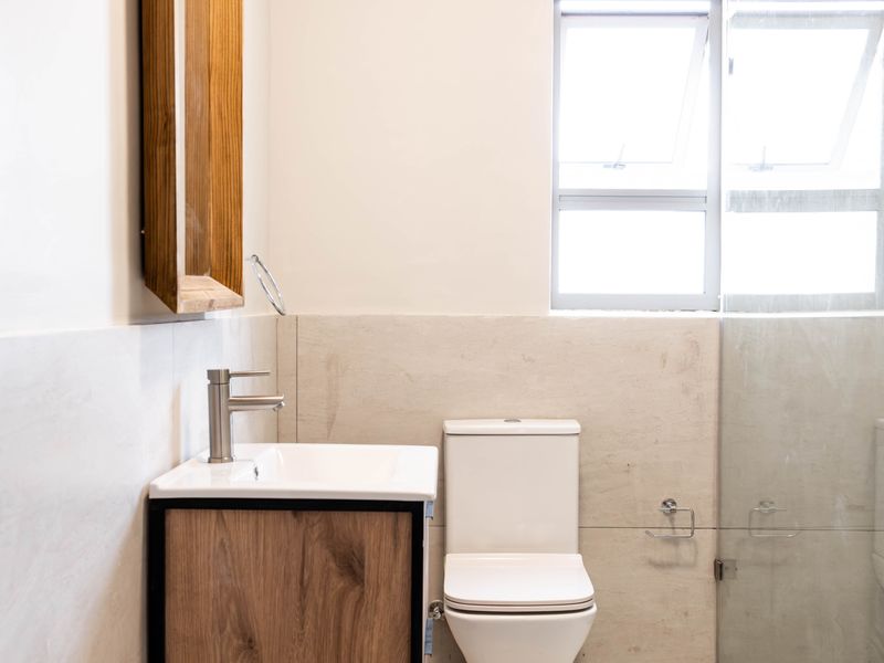 Modern bathroom featuring a wood-finish vanity, white basin, toilet, and a glass-enclosed shower with neutral wall and floor tiling.