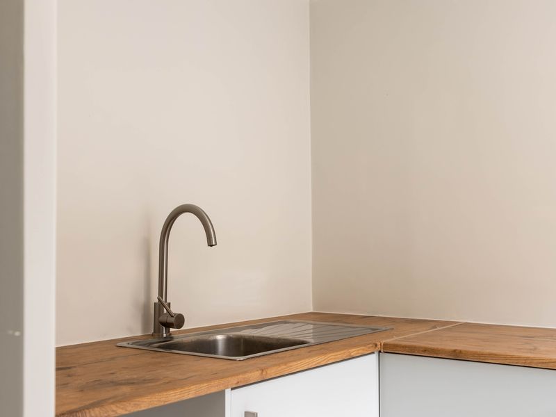 A clean and modern scullery or kitchenette area featuring a stainless steel sink, gooseneck faucet, and timber laminate countertop with white cabinetry.