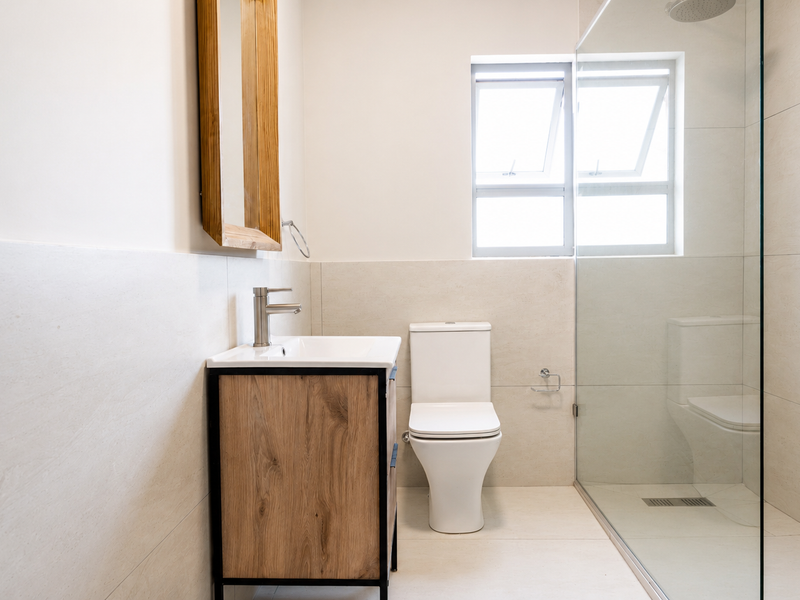 Modern bathroom featuring a wood-finish vanity, white ceramic basin, toilet, and a spacious walk-in shower with a glass enclosure and neutral wall tiling.