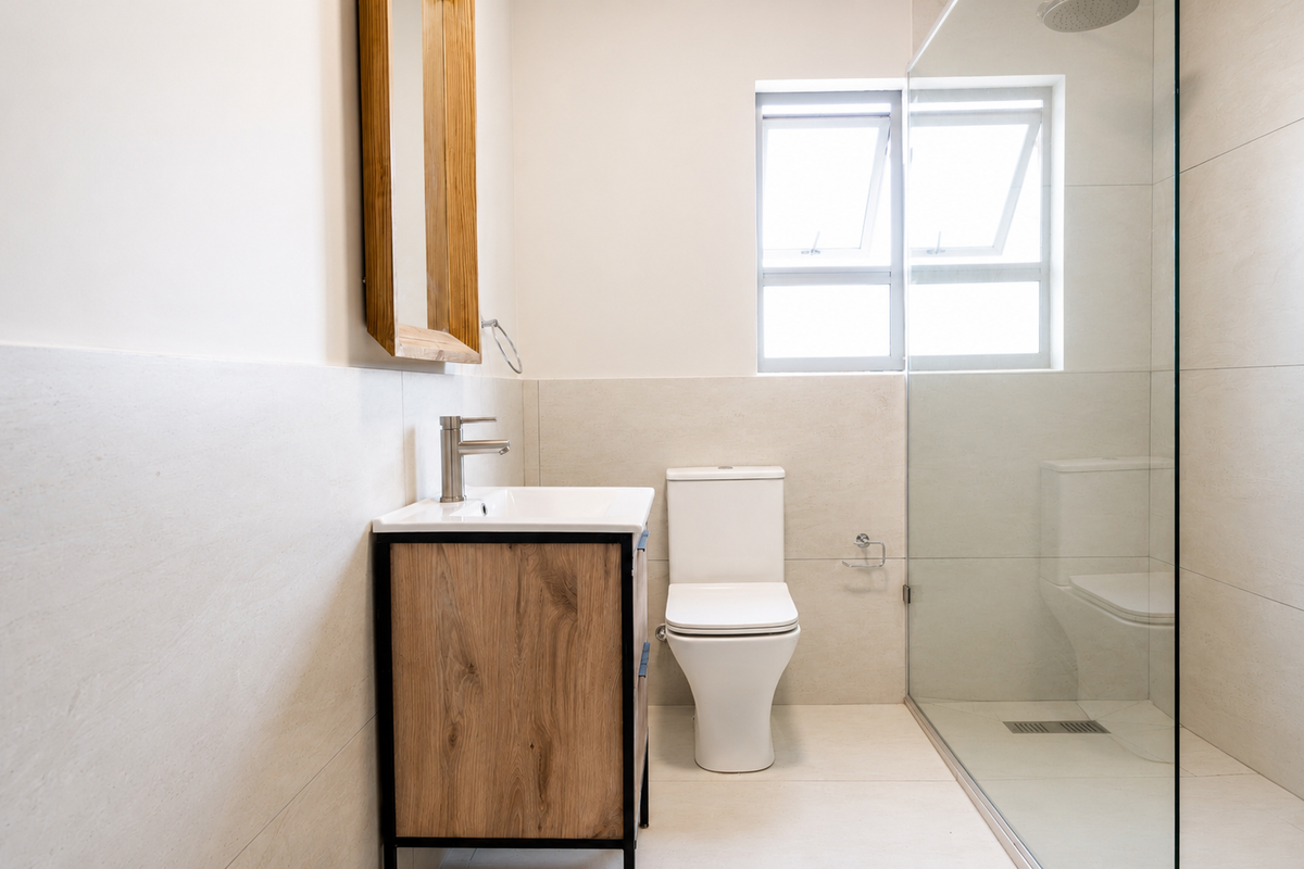 Modern bathroom featuring a wood-finish vanity, white ceramic basin, toilet, and a spacious walk-in shower with a glass enclosure and neutral wall tiling.