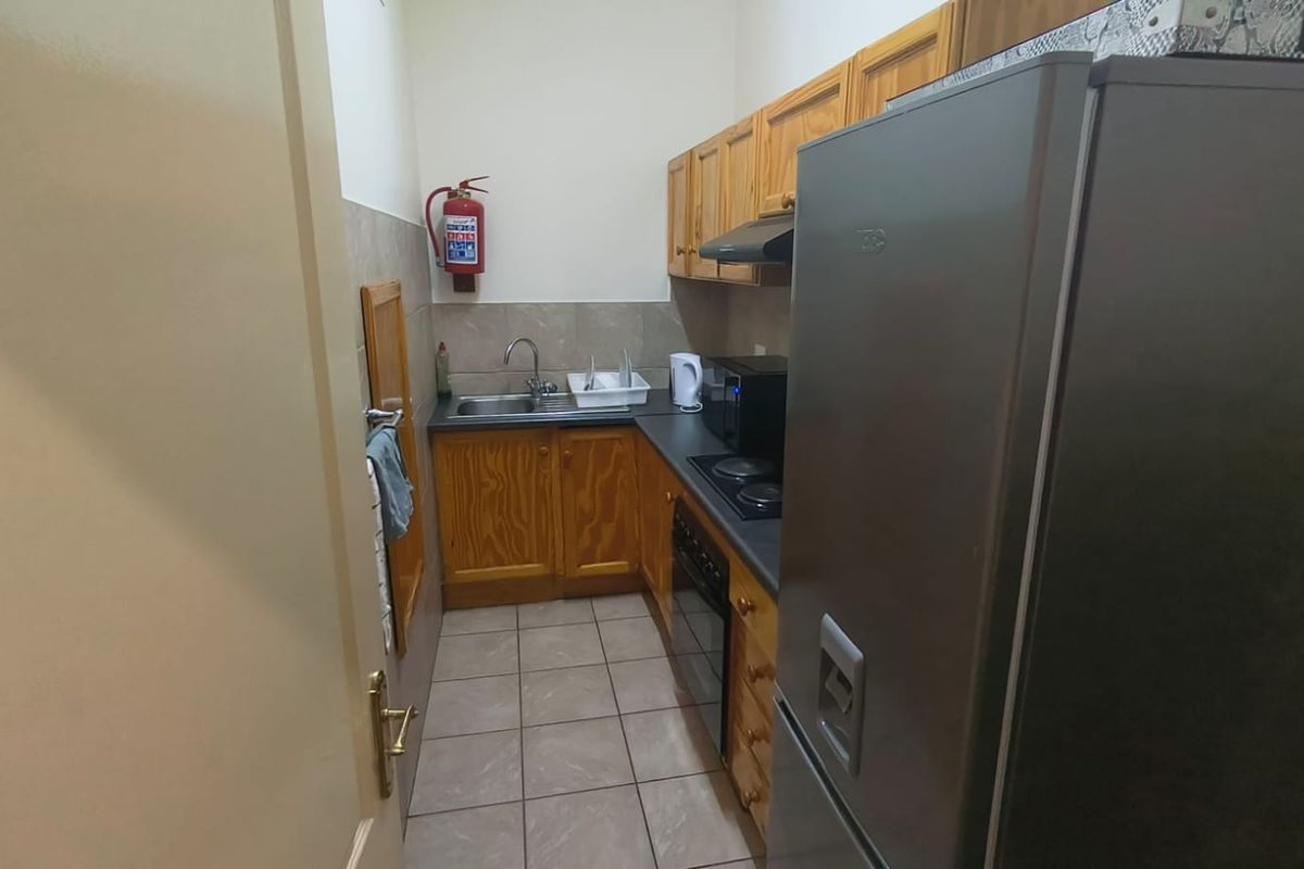 A compact office kitchenette featuring wooden cabinetry, a stainless steel sink, built-in oven, and electric hob, finished with tiled flooring.