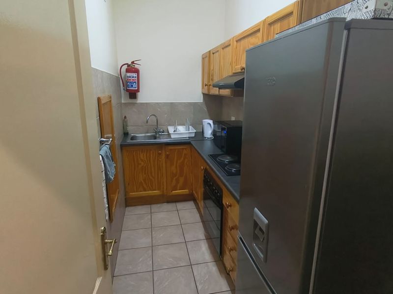 A compact office kitchenette featuring wooden cabinetry, a stainless steel sink, built-in oven, and electric hob, finished with tiled flooring.