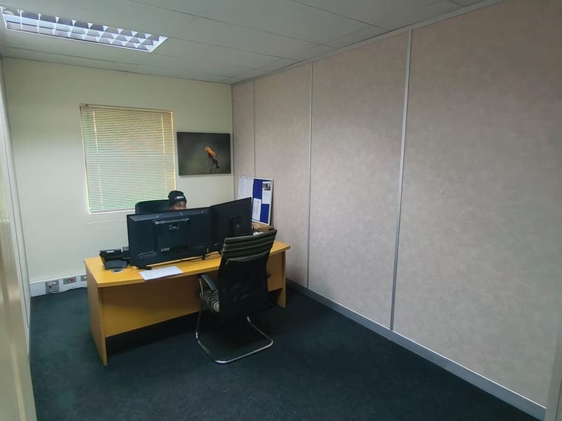 A functional office space featuring modular partition walls, carpeted flooring, a suspended ceiling with recessed lighting, and a window with venetian blinds.