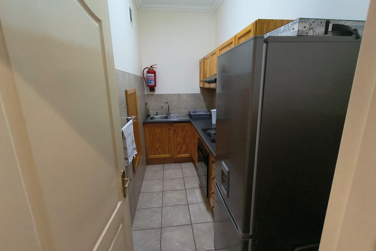 A compact office kitchen featuring wooden cabinetry, tiled flooring, a stainless steel sink, and built-in cooking appliances.