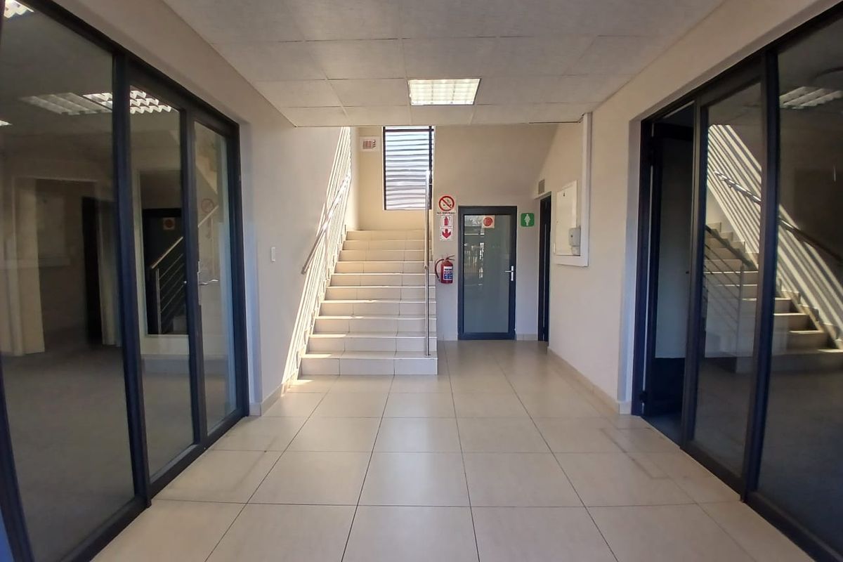 A bright, modern office hallway featuring tiled flooring, a central staircase, and glass-partitioned office entrances with recessed ceiling lighting.