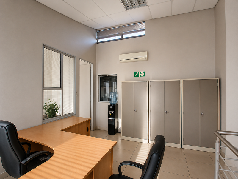 A clean, professional office space featuring tiled flooring, built-in metal storage cabinets, a split-system air conditioner, and ample natural light through aluminium-framed windows.