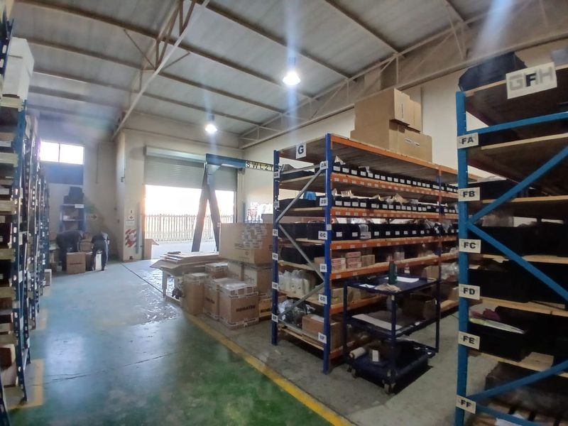 Industrial warehouse space featuring high-bay lighting, exposed steel roof trusses, heavy-duty shelving units, and a large roller shutter door for loading access.