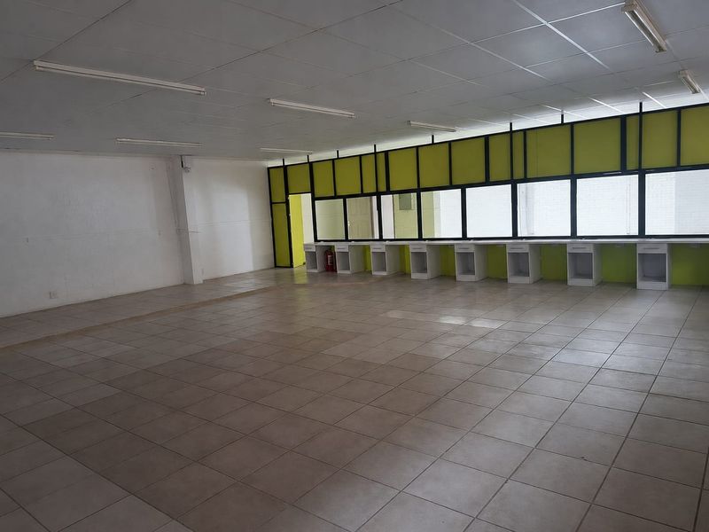 An open-plan commercial office space featuring tiled flooring, a suspended ceiling with fluorescent lighting, and a row of built-in desk units.