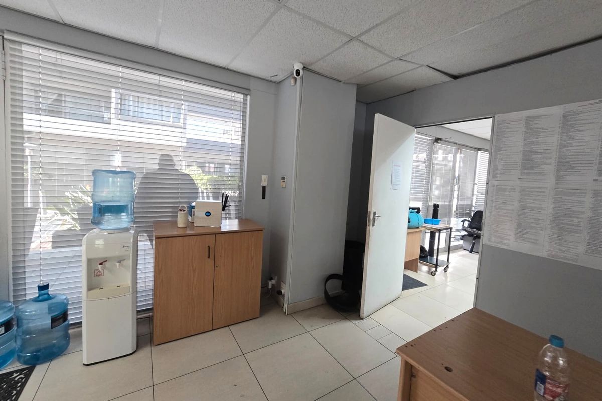 A functional office space featuring tiled flooring, a suspended ceiling, and large windows with venetian blinds, providing natural light and access to adjacent rooms.
