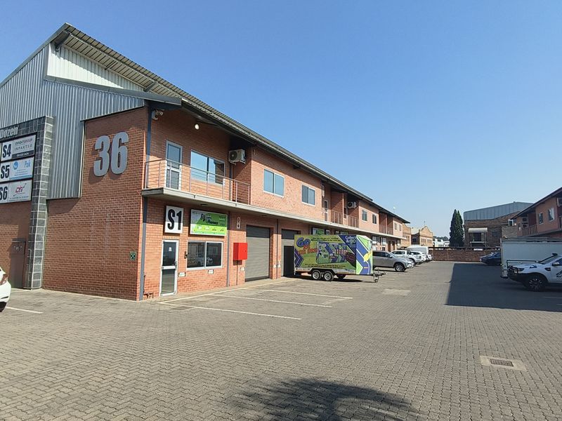 Exterior view of a multi-unit industrial complex featuring brick construction, metal roofing, roller shutter doors, and a paved parking area.