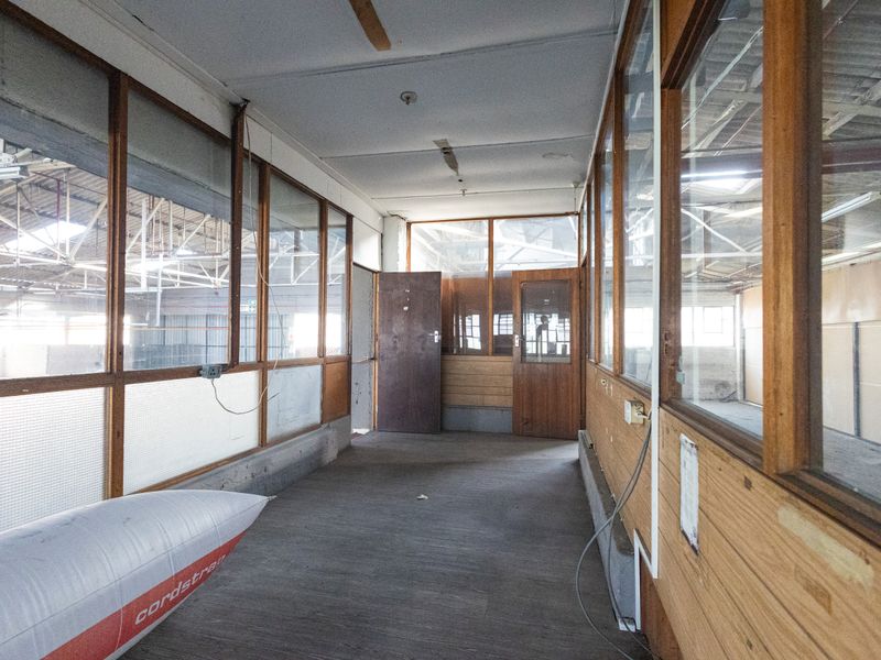 An industrial hallway featuring timber-framed glass partitions overlooking a warehouse space, with exposed roof trusses and functional access doors.