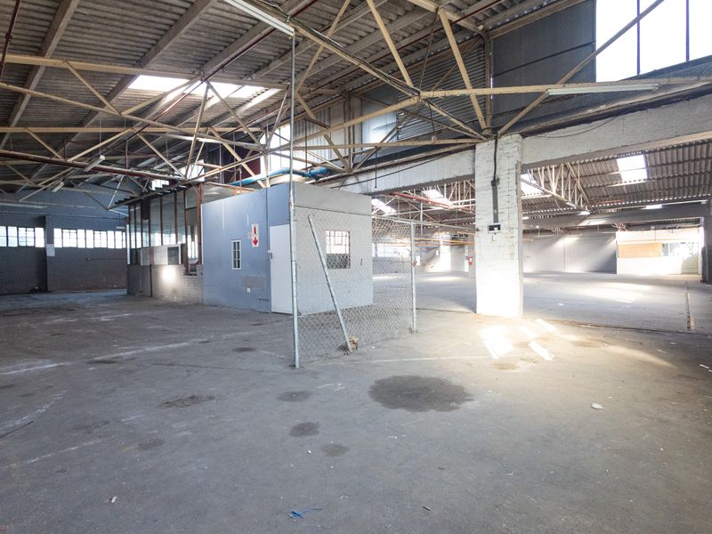 Expansive industrial warehouse space featuring high exposed steel roof trusses, concrete flooring, integrated office enclosure, and natural light from overhead skylights.