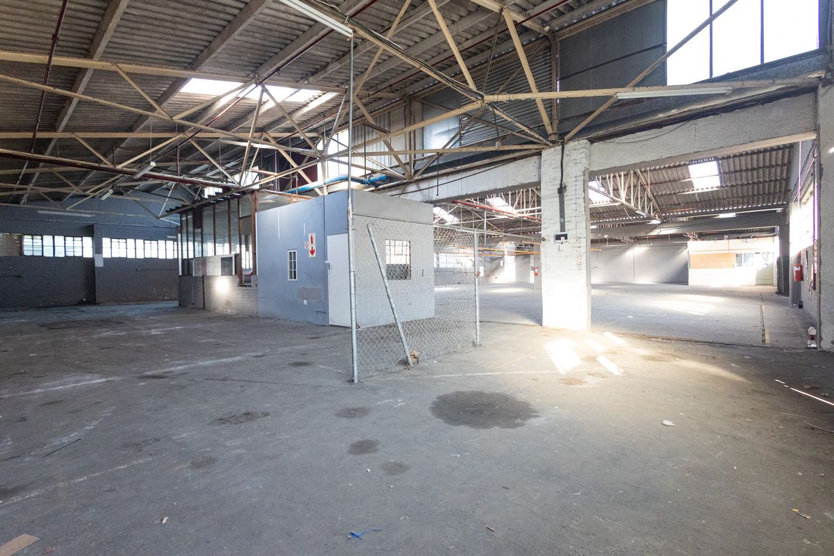 Expansive industrial warehouse space featuring high exposed steel roof trusses, concrete flooring, integrated office enclosure, and natural light from overhead skylights.