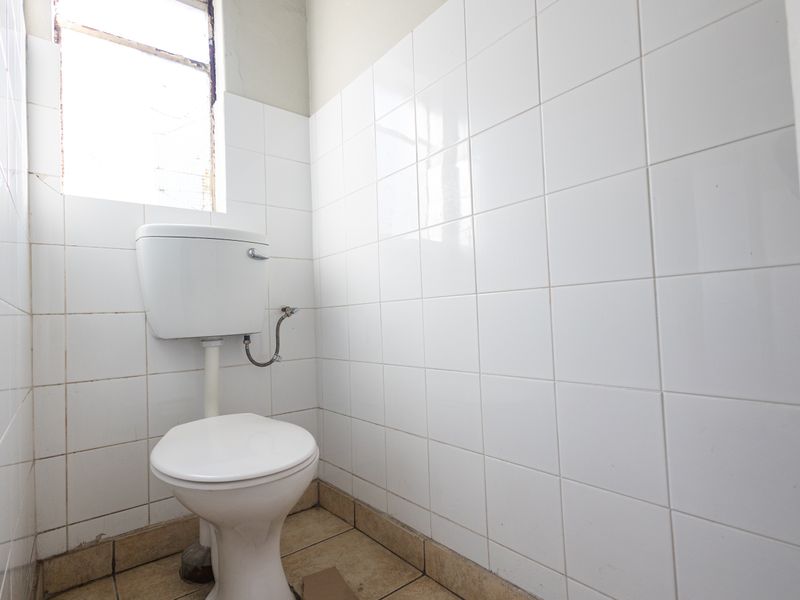 A functional industrial-style bathroom featuring white wall tiles, a standard toilet with a high-level cistern, and tiled flooring.
