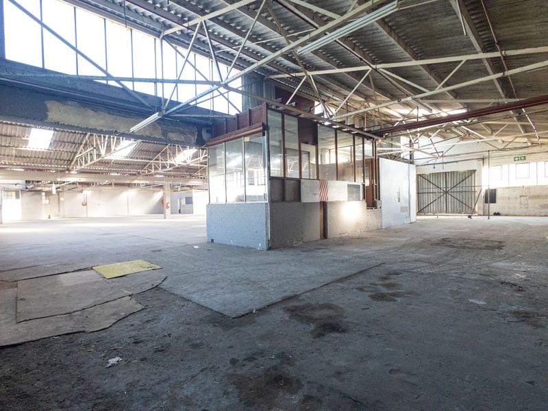 Spacious industrial warehouse featuring high exposed steel roof trusses, concrete flooring, and an internal office enclosure, suitable for various commercial or storage applications.