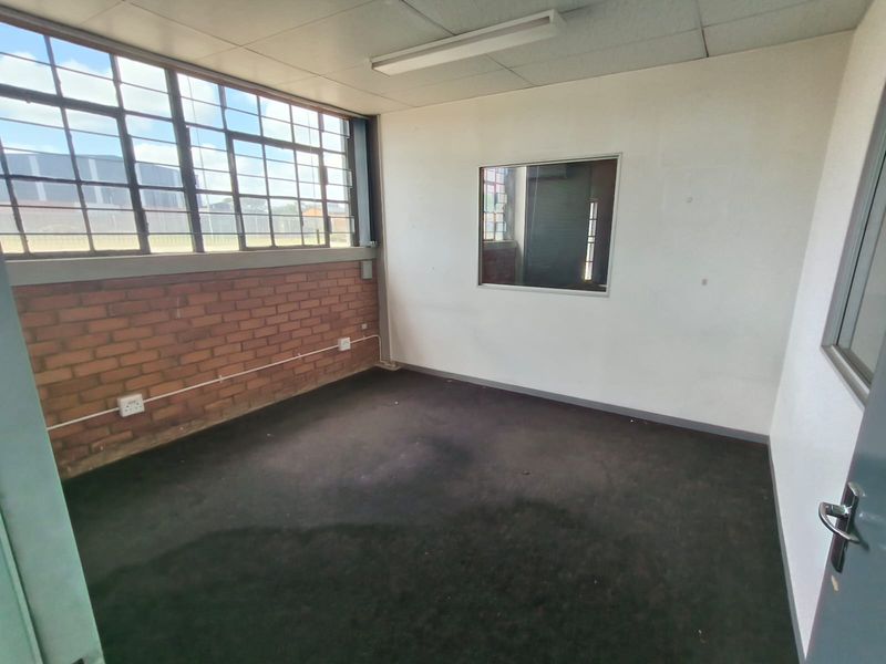Empty industrial office space featuring exposed brickwork, large steel-framed windows, internal viewing pane, and functional overhead lighting.