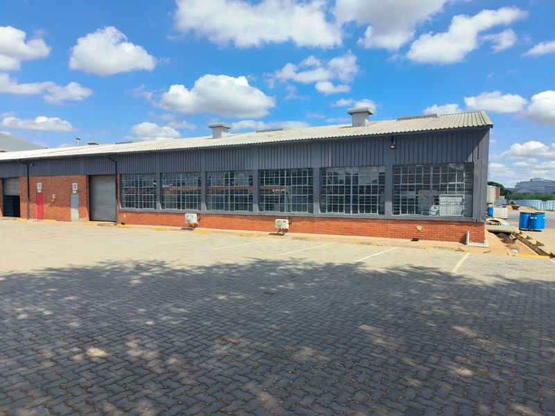 Industrial warehouse exterior featuring brick and metal cladding, large steel-framed windows, roller shutter doors, and a paved parking area.