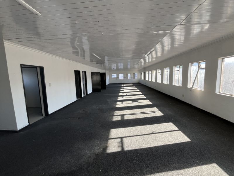 A spacious, empty open-plan office area featuring a suspended ceiling, carpeted flooring, and multiple windows providing natural light.