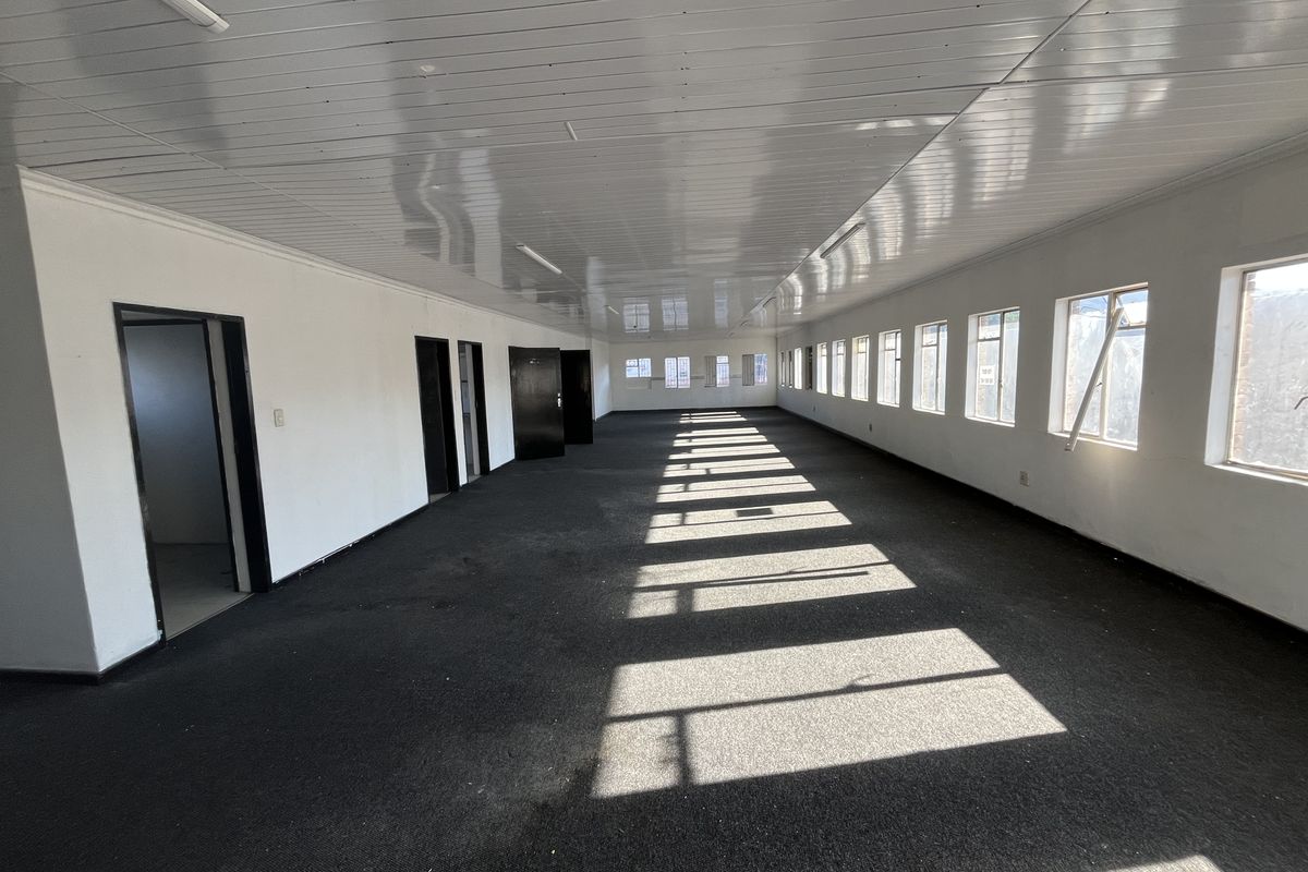 A spacious, empty open-plan office area featuring a suspended ceiling, carpeted flooring, and multiple windows providing natural light.
