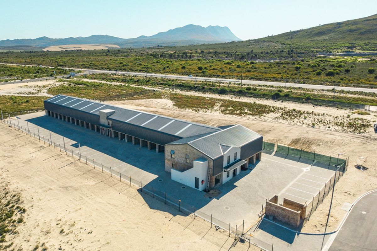 Aerial view of a commercial property featuring a long storage facility with metal roofing, paved parking, and perimeter fencing in a rural landscape.