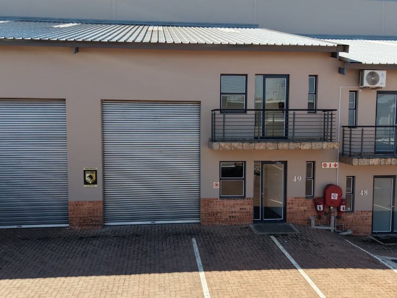 Front elevation of an industrial unit featuring roller shutter doors, a balcony with metal railings, and dedicated paved parking.
