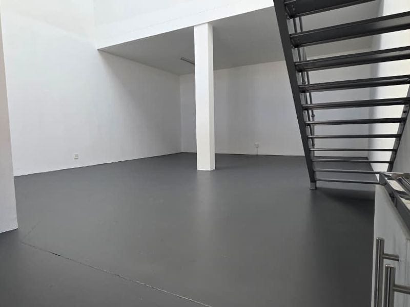 Spacious open-plan industrial unit featuring a modern metal staircase, durable epoxy flooring, and a functional kitchenette area with a sink.
