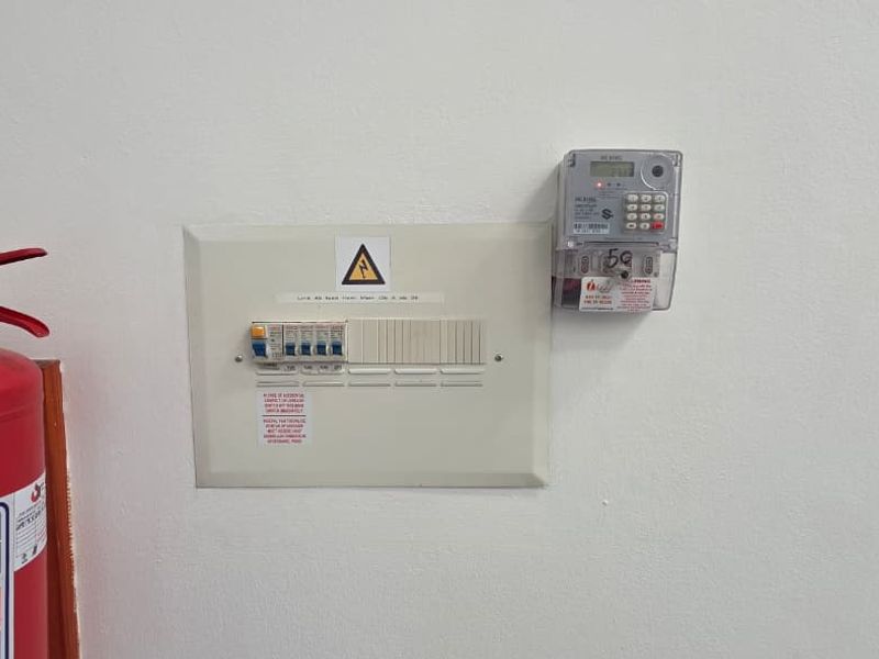 Wall-mounted electrical distribution board and prepaid electricity meter, accompanied by a fire safety extinguisher in a clean utility area.