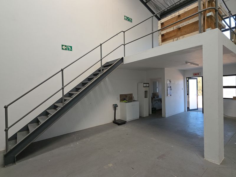 Industrial warehouse space featuring a mezzanine level, metal staircase, roller shutter door, and a small kitchenette area with sink.