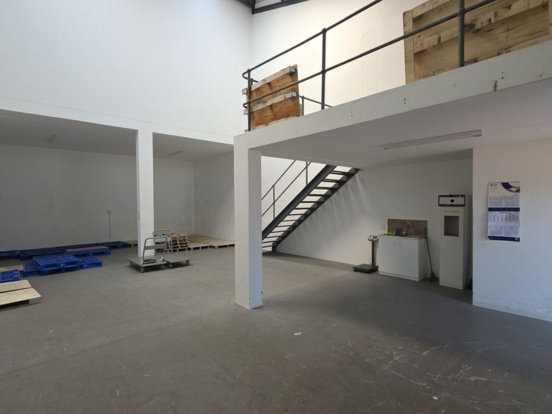 Spacious industrial warehouse unit featuring a mezzanine level, metal staircase, concrete flooring, and a small kitchenette area, suitable for storage or light industrial use.