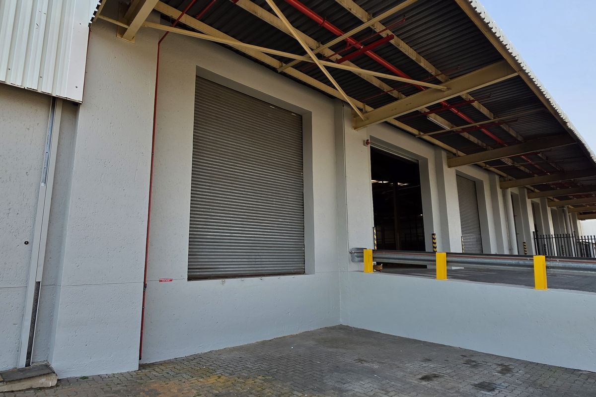 Industrial loading bay area featuring multiple roller shutter doors, a covered canopy, and a paved yard with safety bollards.