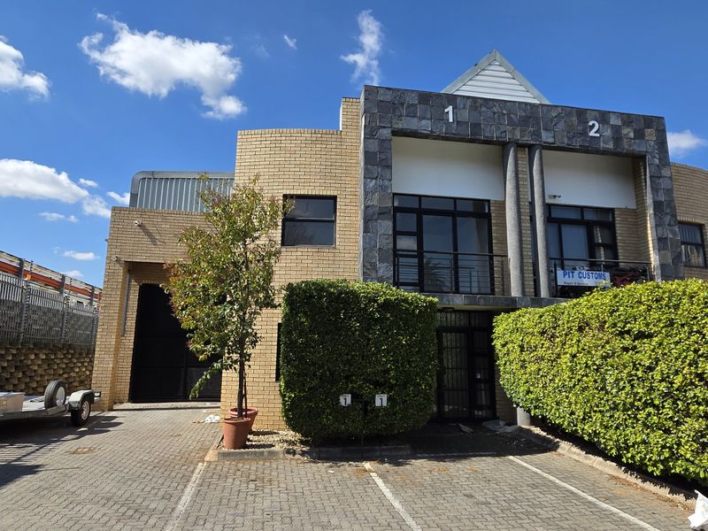 Front elevation of a multi-unit industrial property featuring brick and stone facade, paved parking area, and secure perimeter fencing.