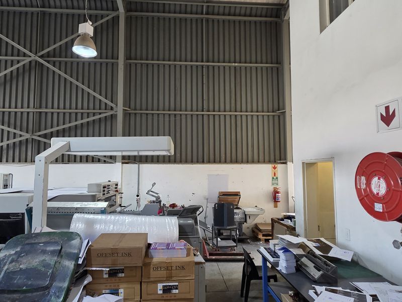 Industrial workspace featuring high-clearance corrugated metal walls, exposed steel roof structure, and essential fire safety equipment including a hose reel and extinguisher.