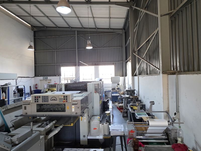 Spacious industrial utility area featuring high ceilings with exposed steel trusses, corrugated metal wall cladding, and concrete flooring, currently utilized for printing operations.
