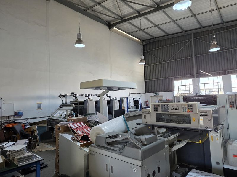 Spacious industrial warehouse area featuring high ceilings, exposed steel roof trusses, large windows for natural light, and functional pendant lighting.