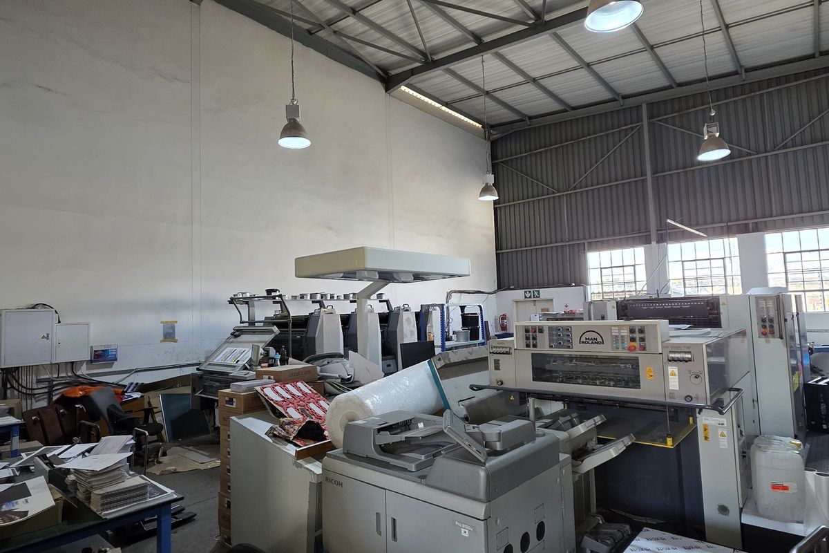 Spacious industrial warehouse area featuring high ceilings, exposed steel roof trusses, large windows for natural light, and functional pendant lighting.