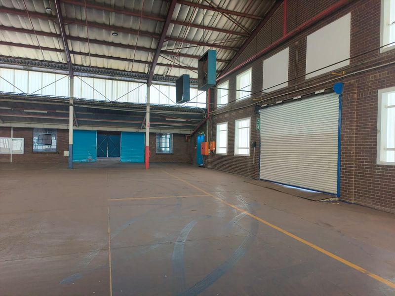 Spacious industrial warehouse featuring high ceilings with exposed steel trusses, concrete flooring, and a large roller shutter door for easy access.