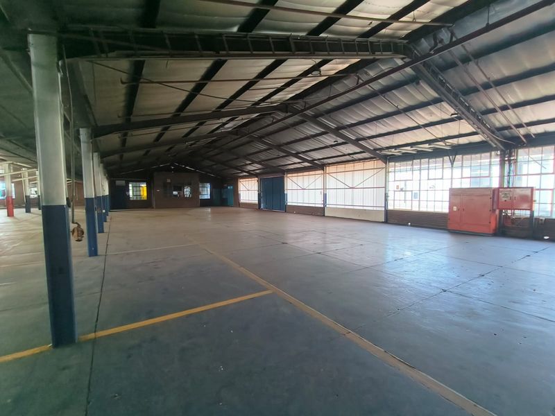 Spacious industrial warehouse area featuring high ceilings with exposed steel trusses, concrete flooring, and large windows providing ample natural light.