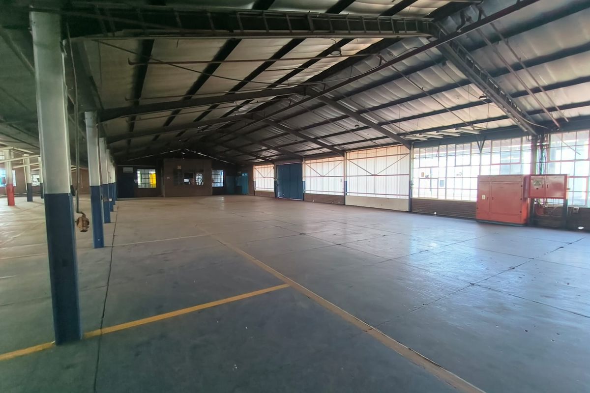 Spacious industrial warehouse area featuring high ceilings with exposed steel trusses, concrete flooring, and large windows providing ample natural light.