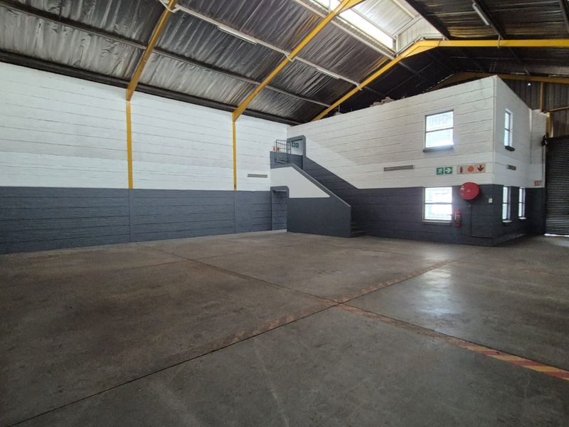 Spacious industrial warehouse unit featuring high ceilings with exposed steel trusses, concrete flooring, a roller shutter door, and an internal mezzanine level with staircase.