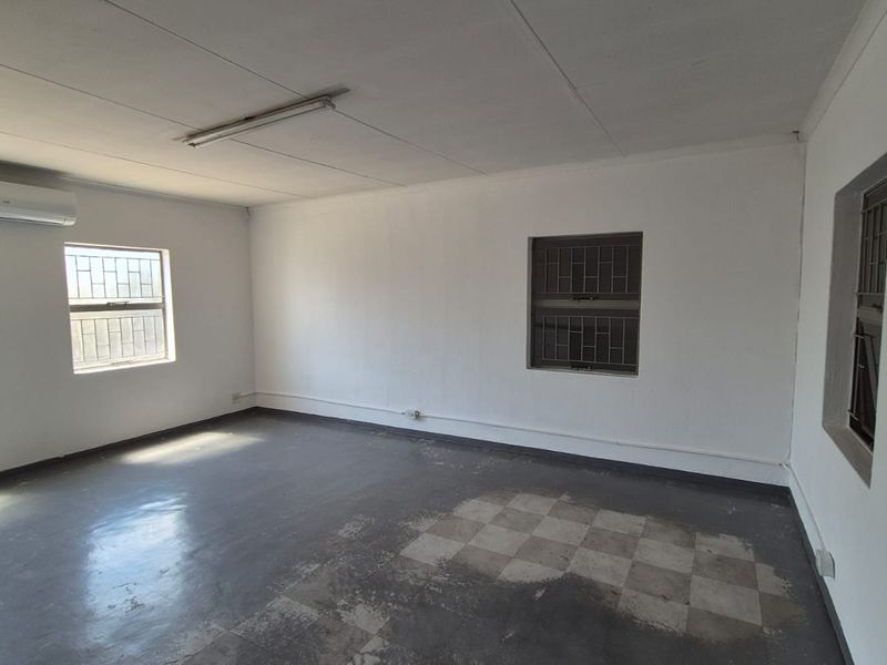An empty, functional office space featuring air conditioning, security bars on windows, and worn tiled flooring, suitable for various industrial or commercial uses.