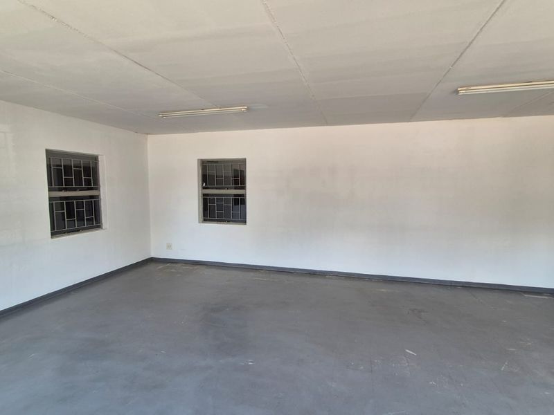 A spacious, empty industrial utility room featuring concrete flooring, fluorescent lighting, and windows fitted with security bars.