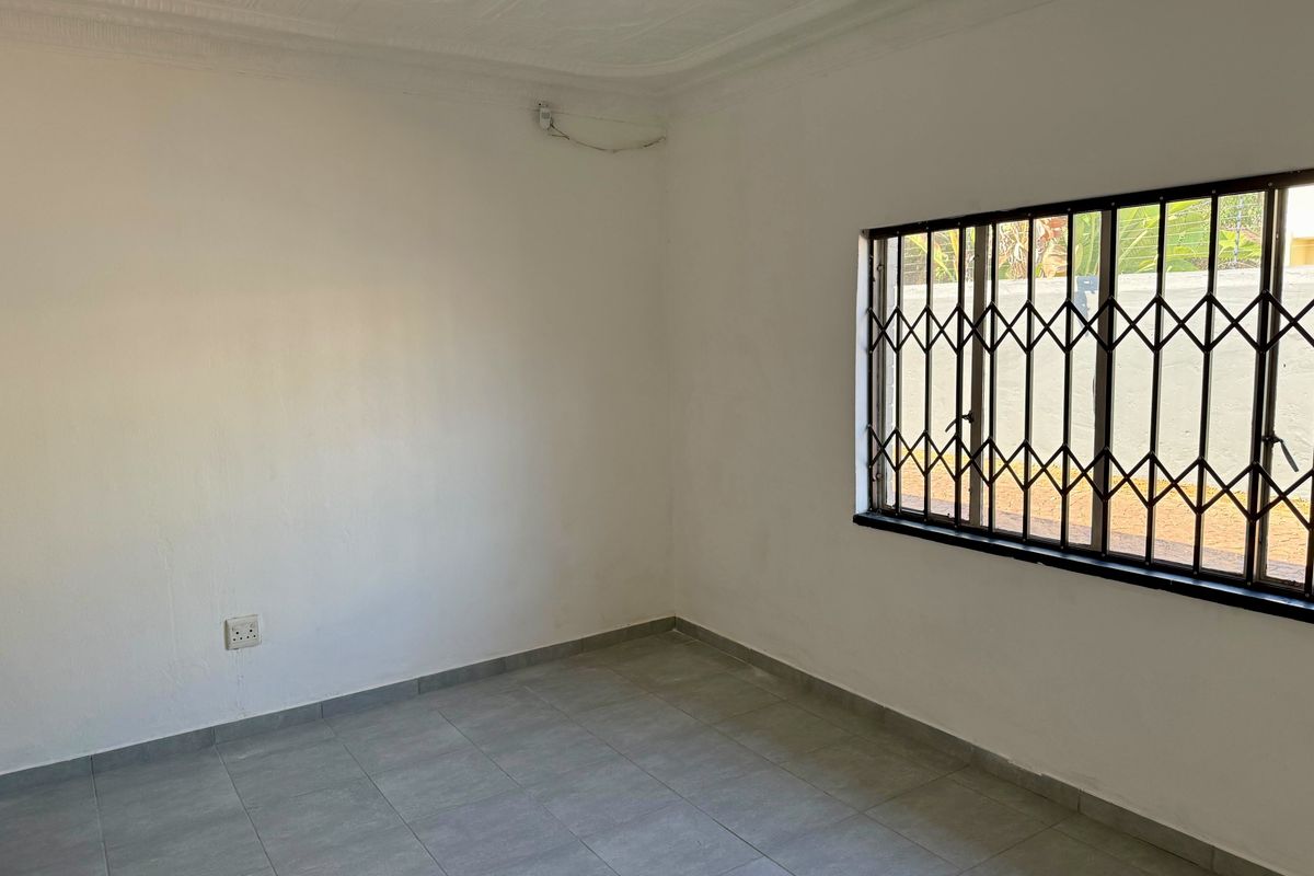 A clean, empty bedroom featuring neutral tiled flooring, a security gate on the window, and decorative ceiling cornicing.
